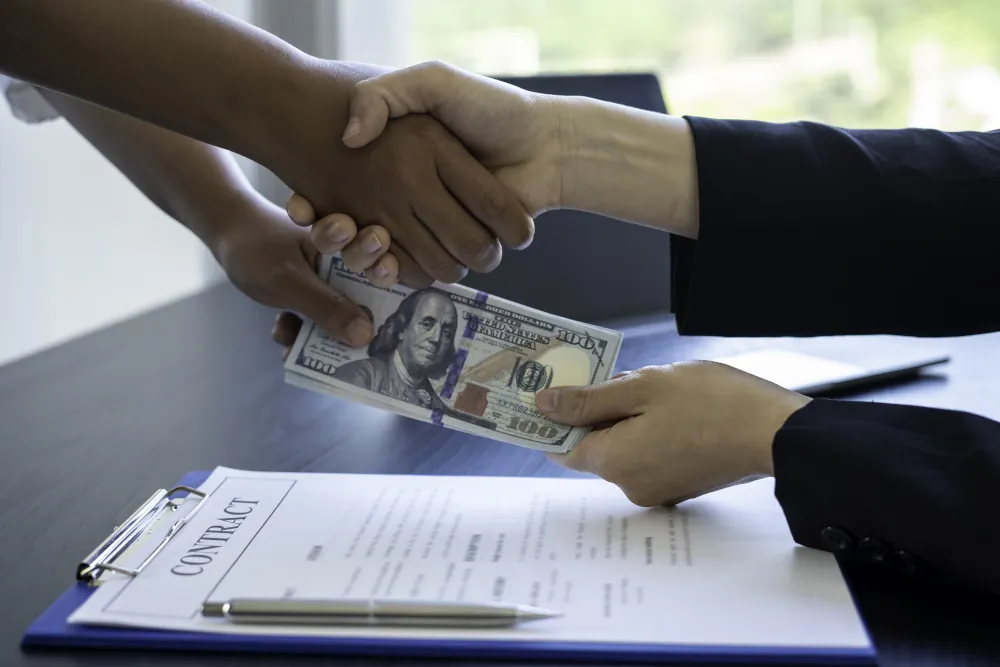 Hands shaking over a contract on a desk. Money is being exchanged at the same time with the other hand.