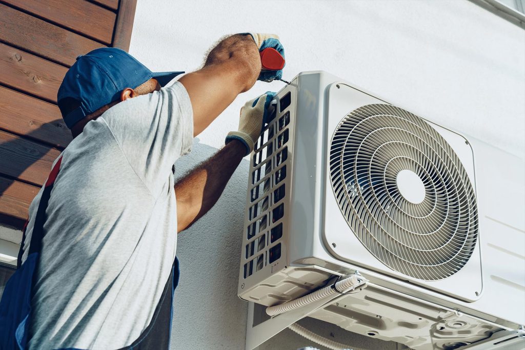 Male technician in overalls and a blue cap repairs an air conditioner on the wall