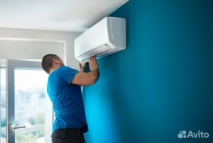 A man in a blue shirt installs a wall-mounted air conditioner in a bright room with a blue accent wall and a window nearby